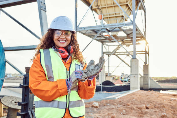 mine manager1 A young African woman mine worker wearing protective wear is looking at the camera while putting on a pair of gloves with coal mine equipment in the background